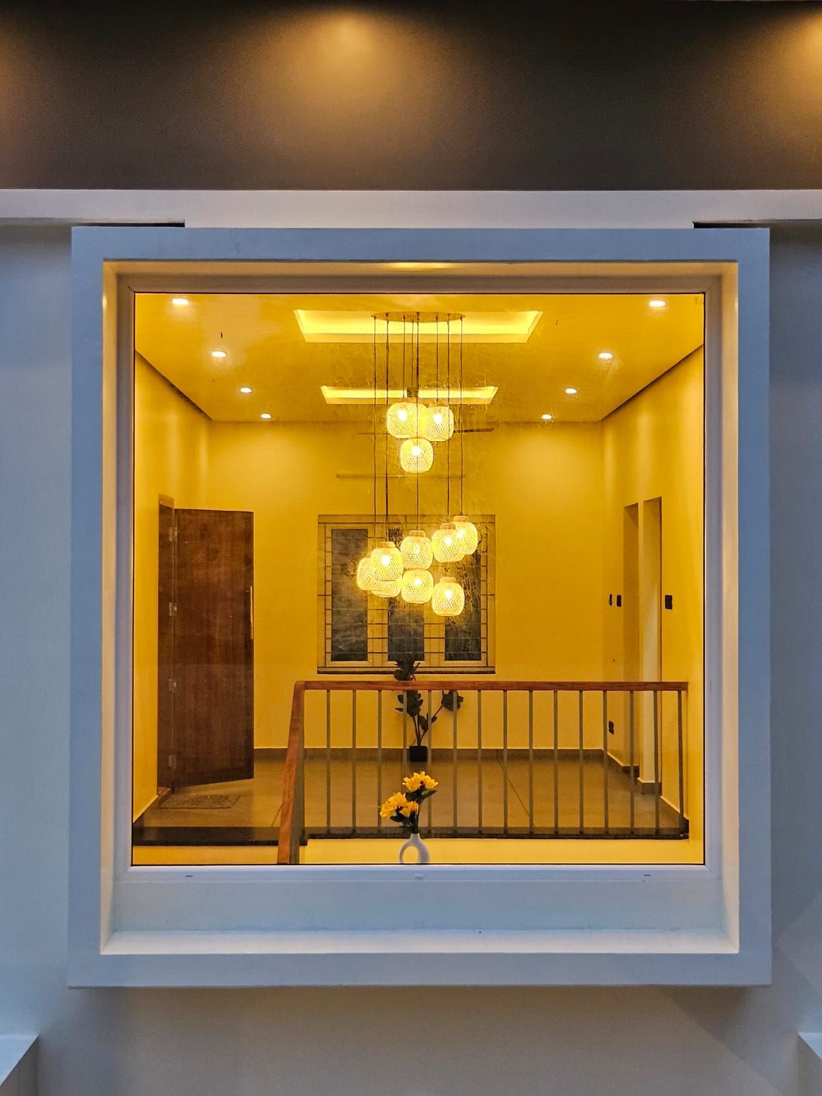 View through a large rectangular window framing an interior staircase landing illuminated by a cluster of warm pendant lights.