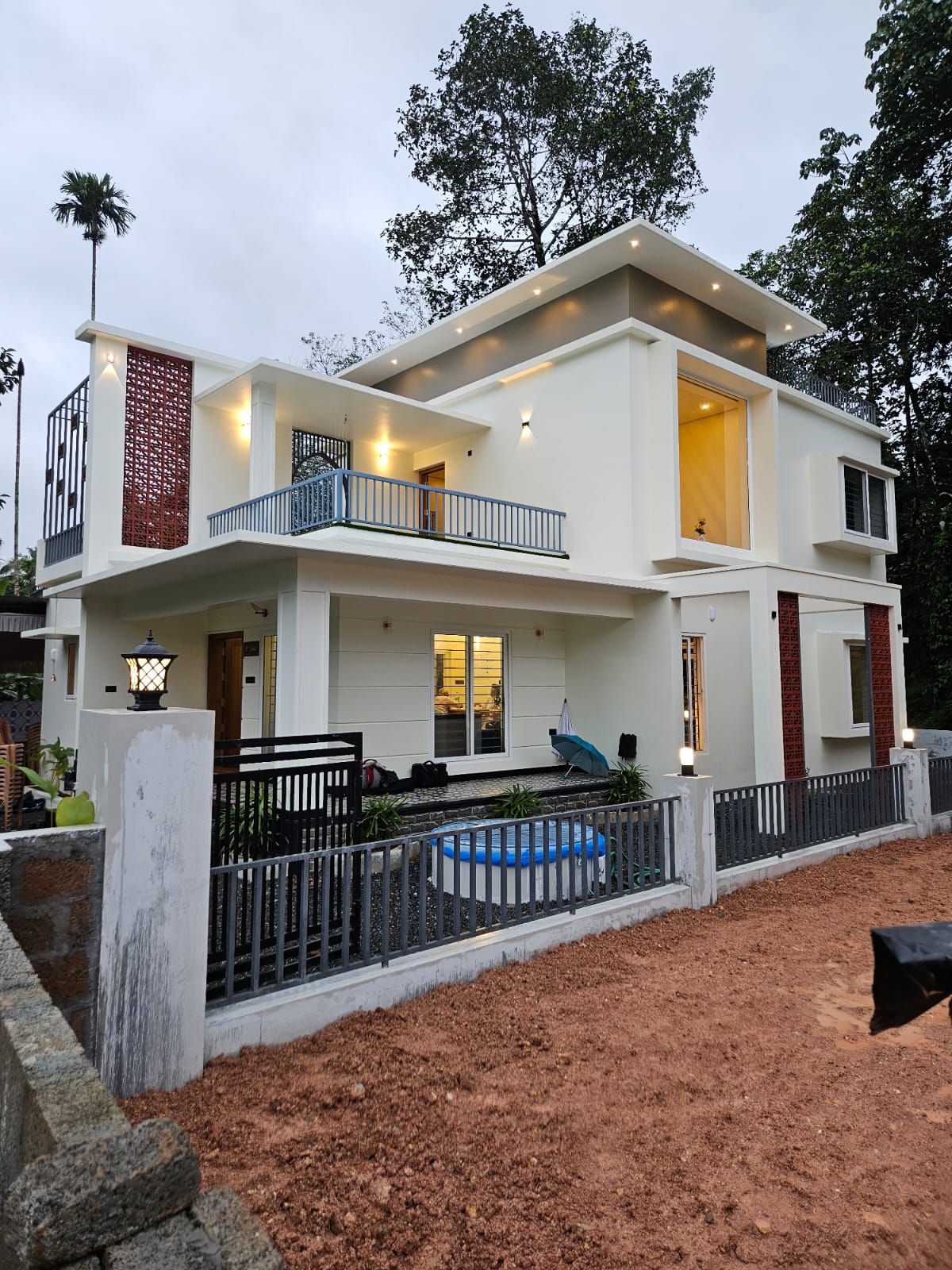 Exterior twilight view of a contemporary two-story white home featuring a flat roof, terracotta breeze blocks, and large illuminated windows set against a tropical backdrop.